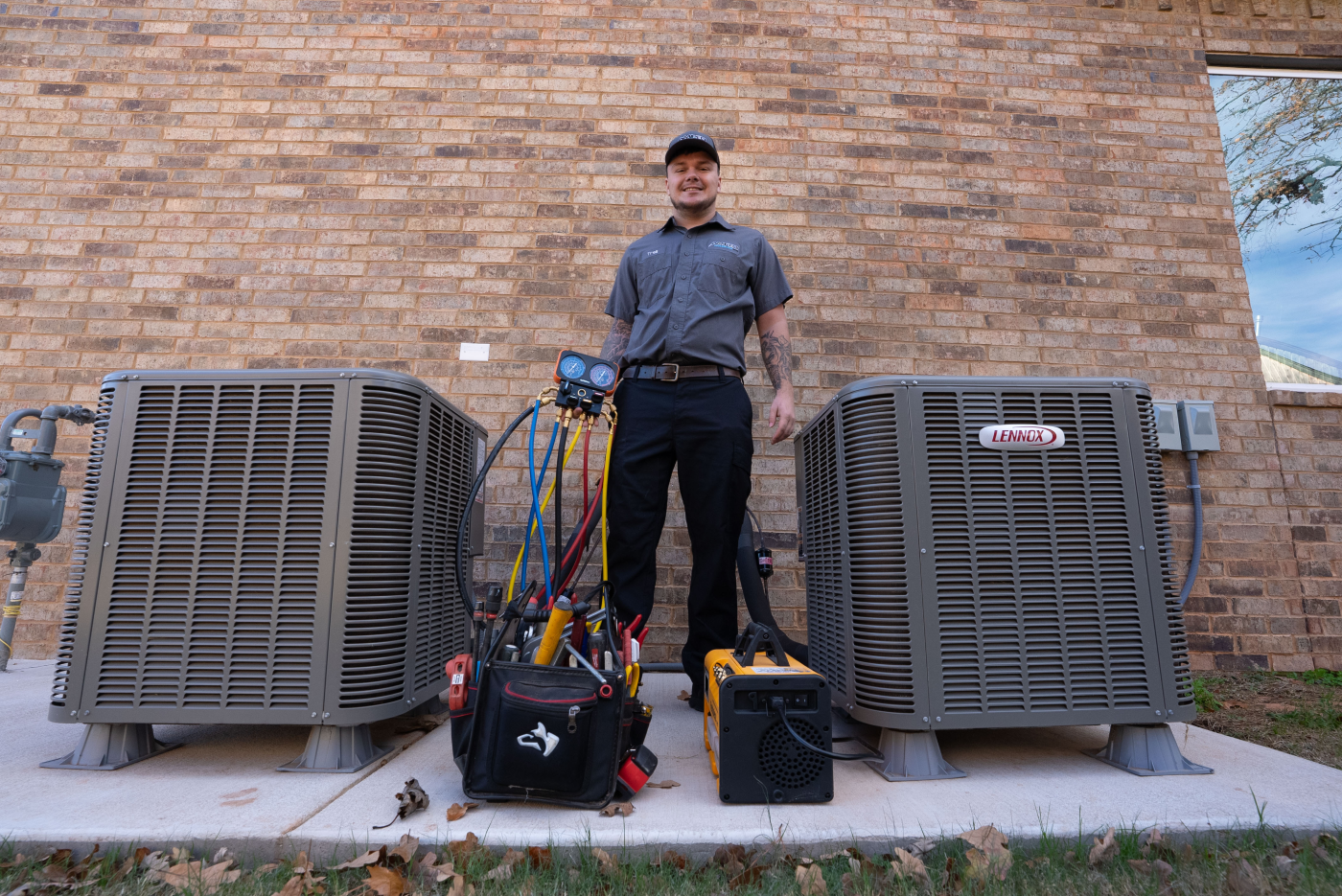 Amped technician stands smiling between two Lennox AC units, with gauges and tool bags placed on the concrete pad below.