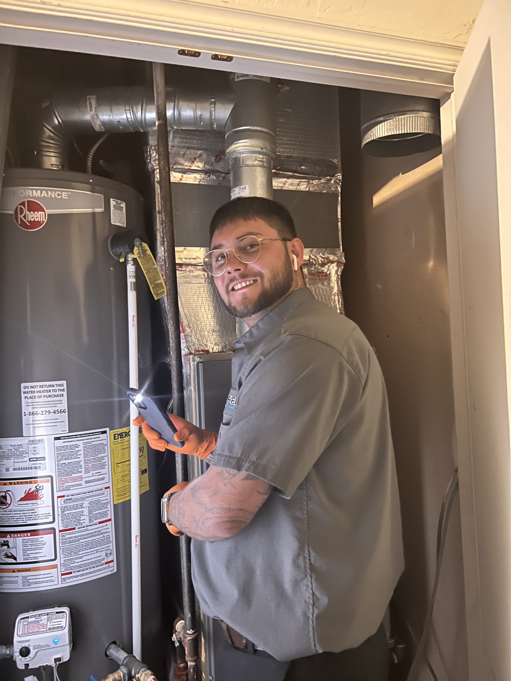 Plumber smiles while inspecting a water heater with a flashlight in a tight utility closet.