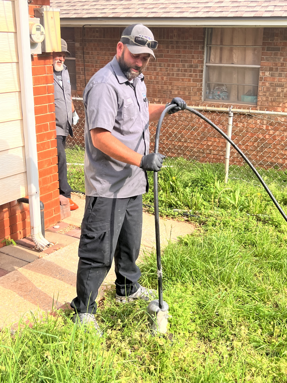 Plumber feeding cable into the ground using drain machine, standing in overgrown yard next to red brick home.