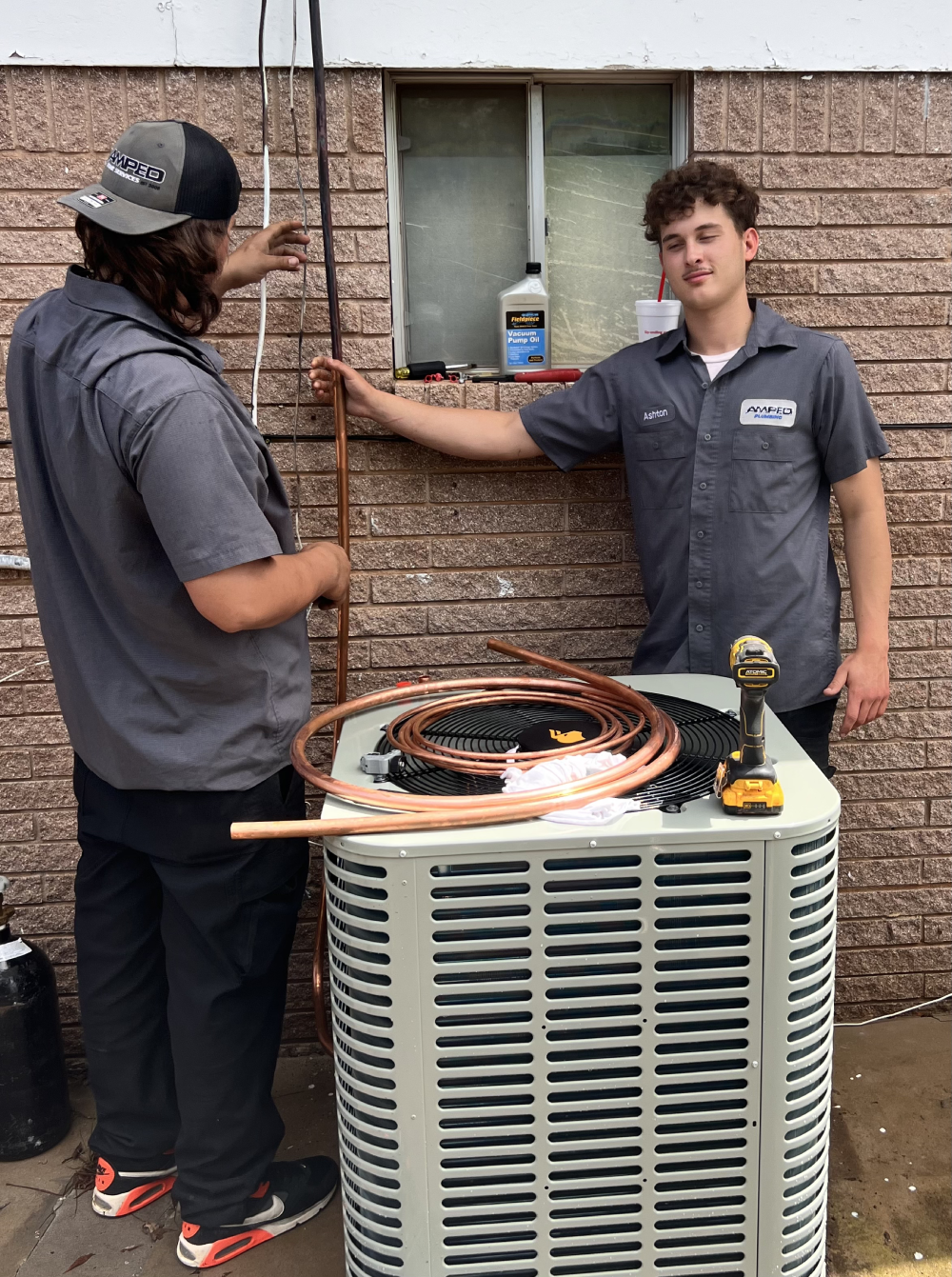 Two HVAC techsare prepping copper lines at the outdoor unit, standing by a brick wall with tools and coiled tubing.