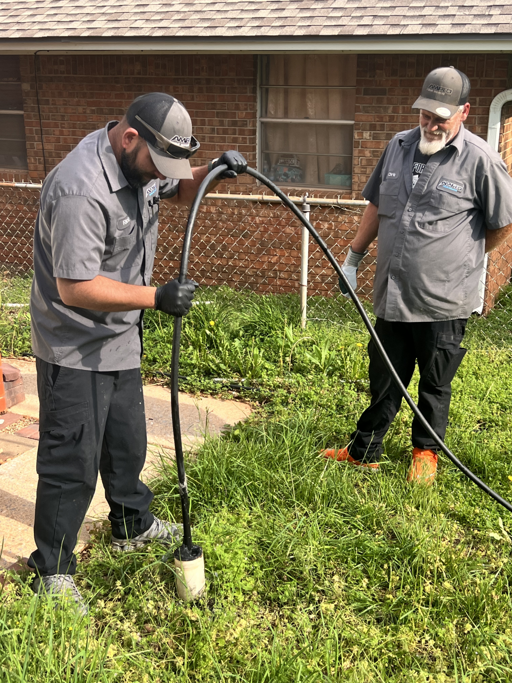 Plumbers working on a pipe in a grassy yard; one inserts equipment into the ground while the other observes.