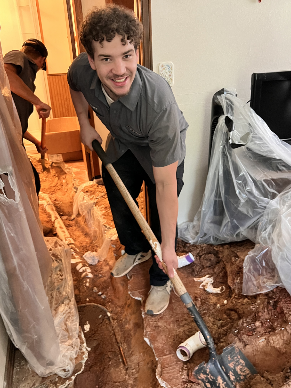 Smiling Plumber digging indoors with a shovel during a plumbing job, surrounded by protective plastic sheets.
