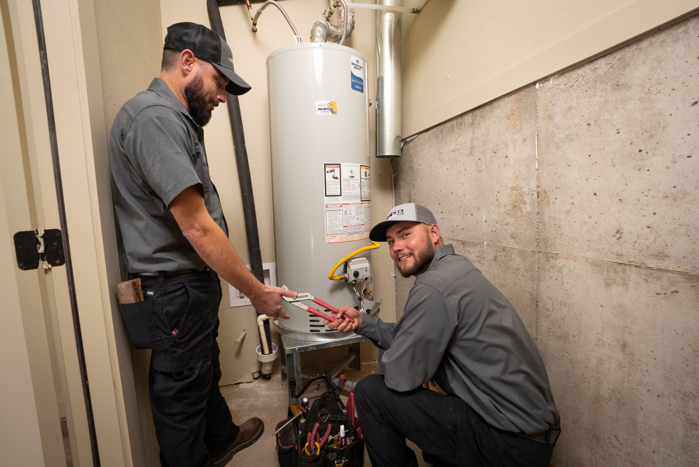Two plumbers techs work on a water heater hookup, one kneeling and smiling while the other passes a pipe wrench.
