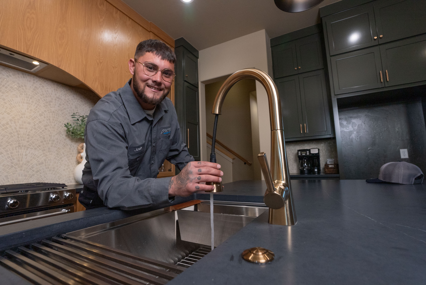 Plumber smiling while testing a modern kitchen faucet, standing behind a black countertop and gold fixture.
