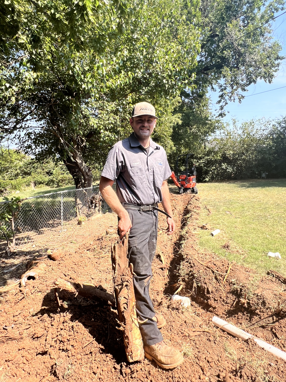 Plumber stands in a sunny yard holding a large tree root beside a trench with exposed pipework.
