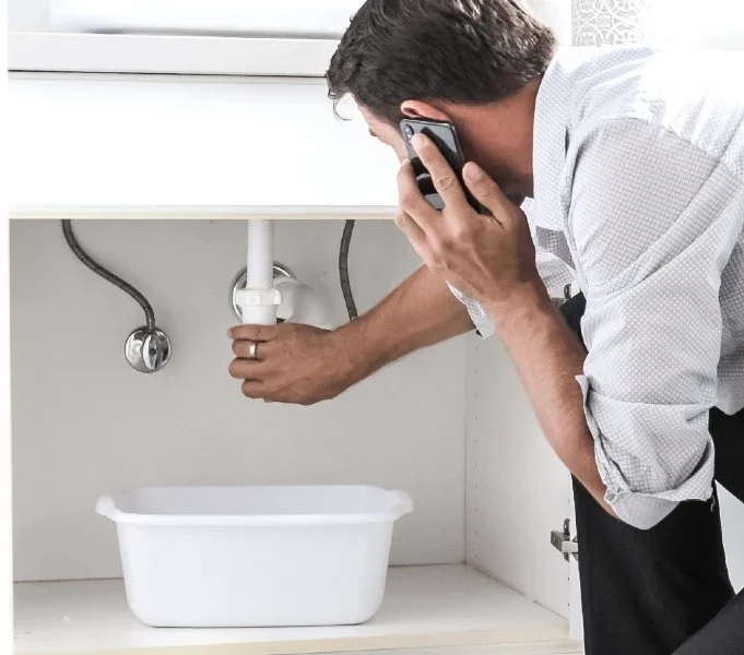 Homeowner checking plumbing under a sink while calling for professional help