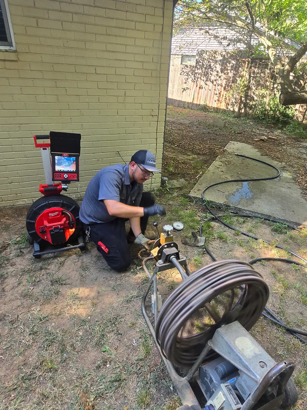 Amped technician using drain-cleaning and sewer-camera equipment beside a brick home