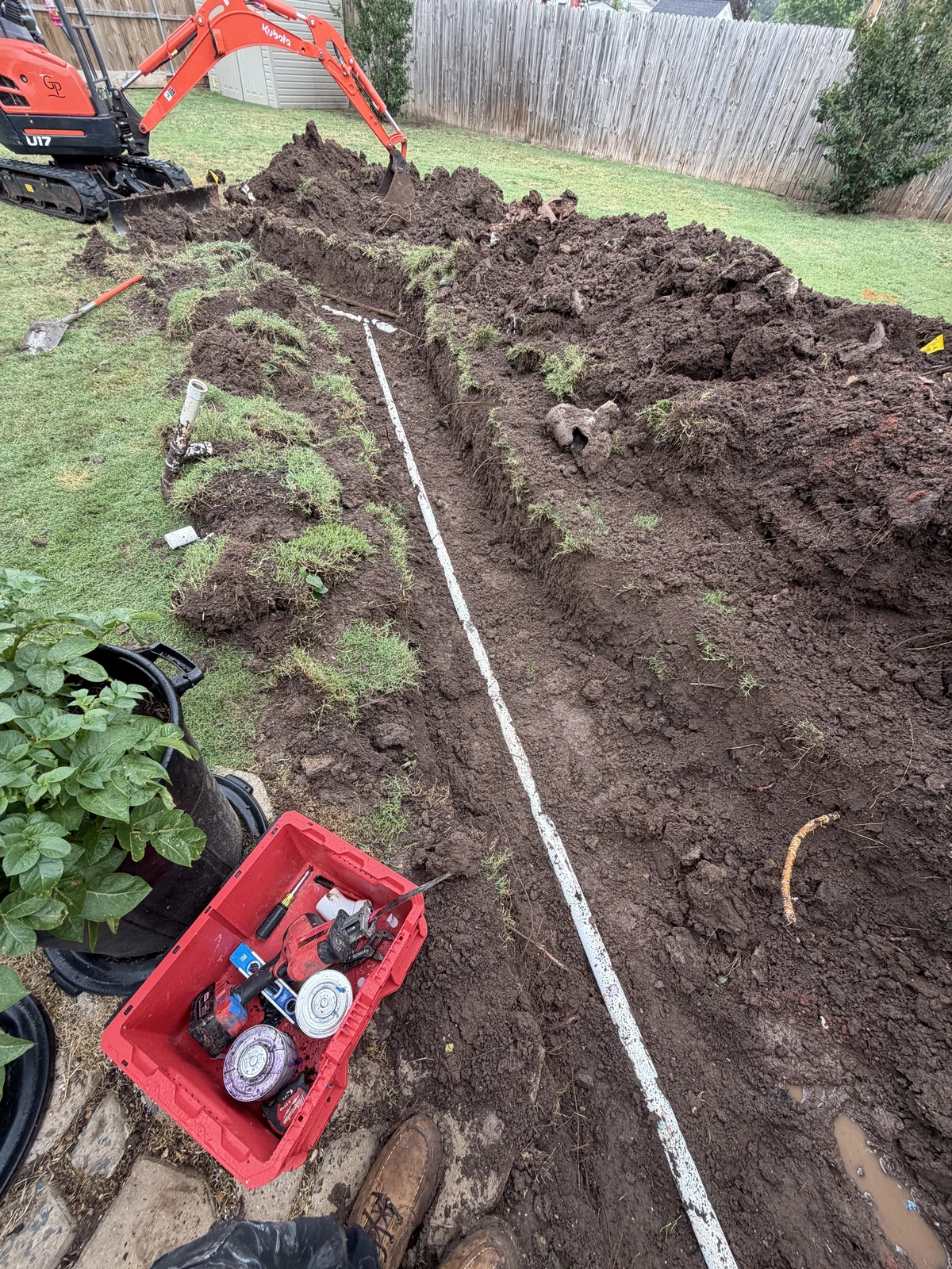 Open yard sewer trench with exposed white piping and a compact excavator