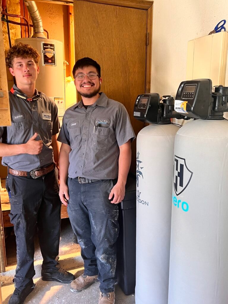 Two Amped Home Services technicians standing beside residential water filtration and softener systems.